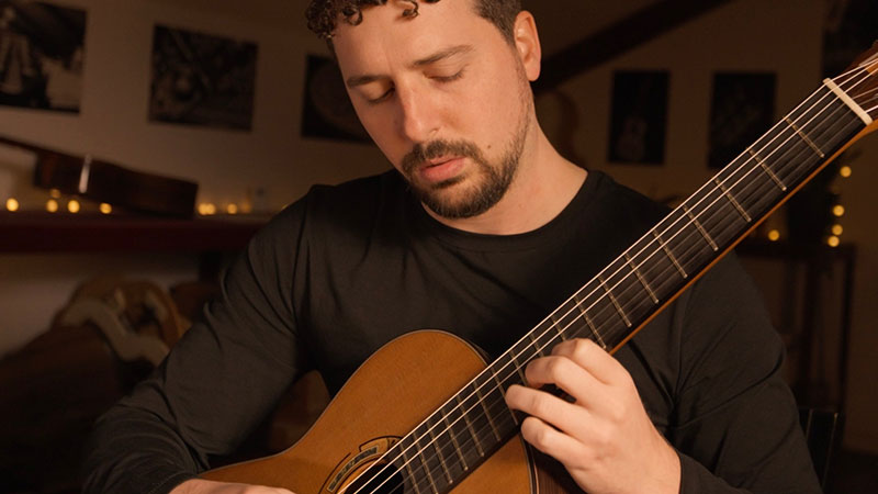 a young white male with short brown hair and short beard and mustache wearing a long sleeved black t shirt playing a guitar