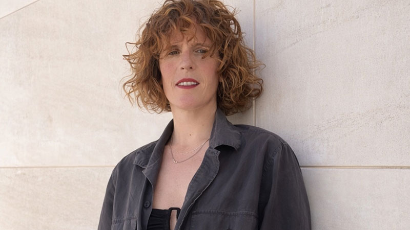 A middle aged white woman with short curly auburn hair wearing red lipstick and a dark blue shirt against a limestone wall