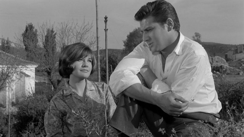 Still from a black and white movie showing a young woman with short hair smiling up at a young white man with short brown swept back hair who is sitting on a wall.