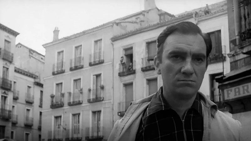 Still from a black and white movie from the mid 1900s era showing a middle-aged white man with short black hair wearing a checked shirt and a light jacket coming toward the camera only showing his head and shoulders, with white buildings in the background