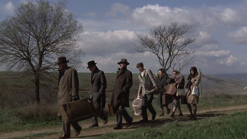 country scene with 4 men and 3 women in mid-1900s clothing walking in a line along a country path