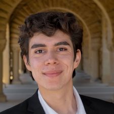 Ramon Salado: young, smiling, white male with short brown curly hair, dark, full eyebrows wearing black suit and white shirt.