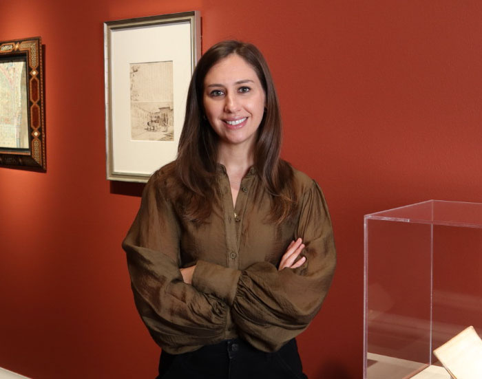CristinaAldrich_Headshot_700x550 Cristina Aldrich: a young woman with long brown hair parted in the middle wearing a brown shirt, crossed arms standing against red wall